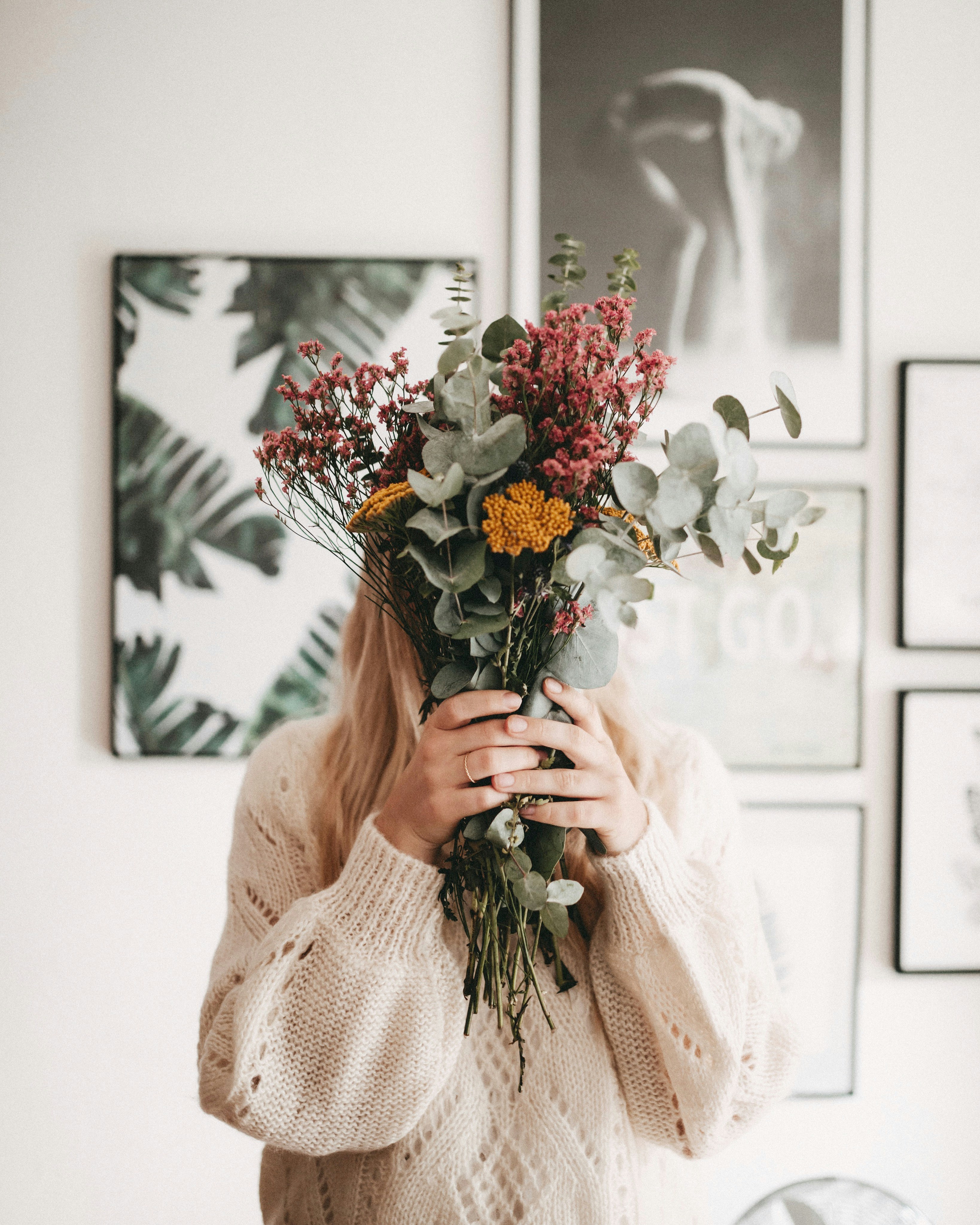 A woman wearing a cream knit sweater holding a beautiful bouquet of mixed flowers in front of her face, standing in a room with nature-themed wall art in the background.