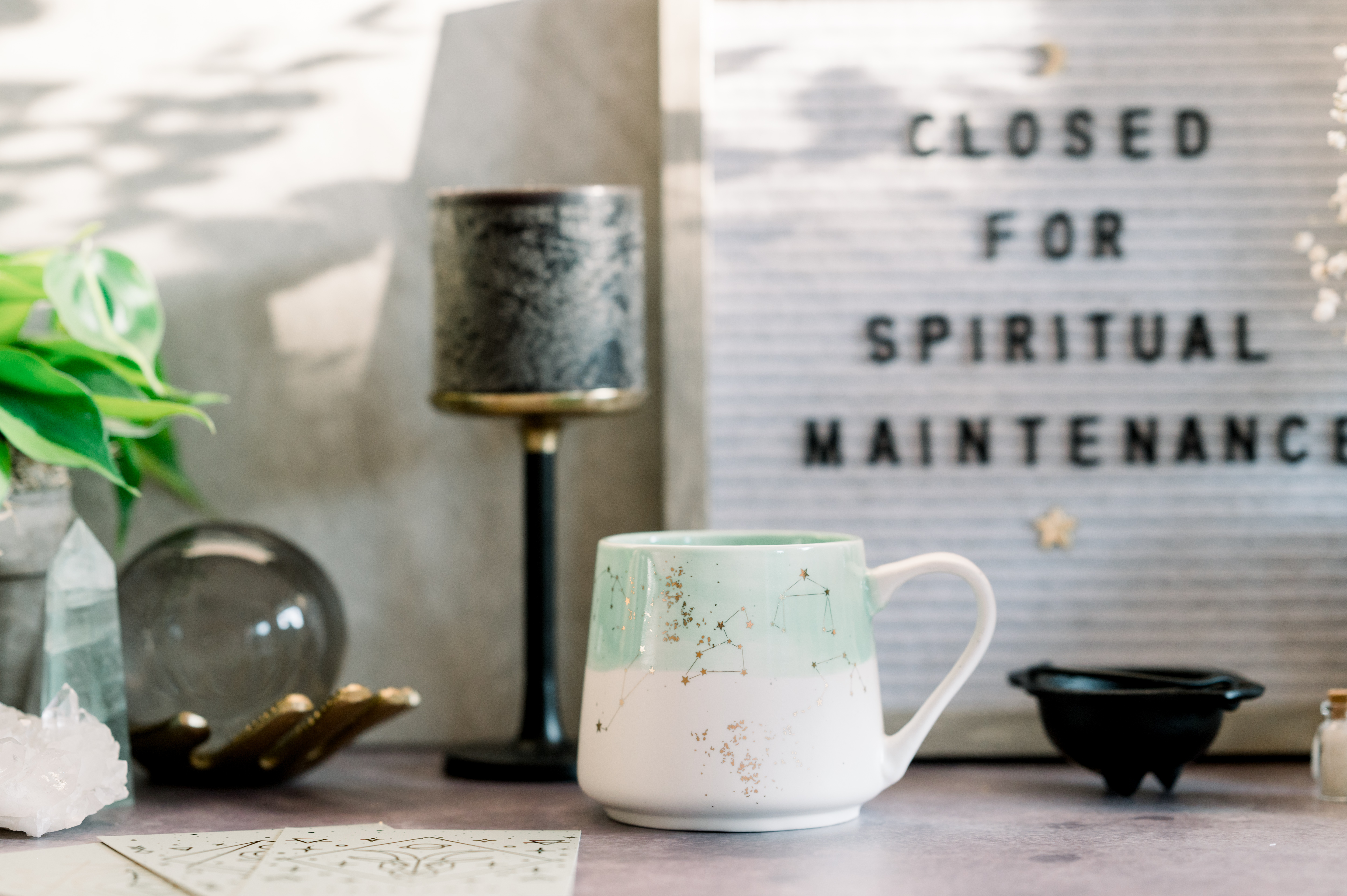 A desk or shelf filled with spiritual items including a mug, crystals, a scrying ball, a candle holder, and a letter board displaying the message 'CLOSED FOR SPIRITUAL MAINTENANCE'.