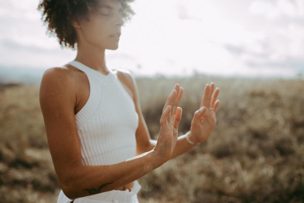 A person with curly hair stands in a field with eyes closed and hands raised, palms forward, as if in meditation or guiding energy. Soft sunlight filters from the background. A person with curly hair stands in a field with eyes closed and hands raised, palms forward, as if in meditation or guiding energy. Soft sunlight filters from the background.