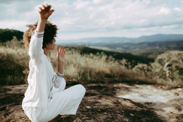 A person with curly hair kneels on a rocky outcrop in a mountainous landscape, hands raised in an upward, receptive gesture, looking away from the viewer.