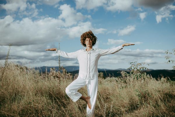 A person with curly hair stands in a tree pose amidst tall grass and rolling hills under a partly cloudy sky, arms extended in balance.