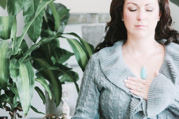 A person with long dark hair, wearing a gray knitted sweater and a turquoise pendant, meditates with closed eyes and one hand gently placed over their heart. A large green plant is visible to the left. A person with long dark hair, wearing a gray knitted sweater and a turquoise pendant, meditates with closed eyes and one hand gently placed over their heart. A large green plant is visible to the left.