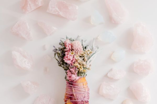 An overhead flat lay of a decorative dried floral and sage smudge stick wrapped with pink string, surrounded by various sizes of raw pink rose quartz crystals and smooth white opalite stones on a light background.