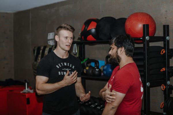 A physiotherapist at Between the Bumpers, a gym in Stittsville, helps a client understand the impact of the stretches he's given him on the client's rehabilitation from his injury. The gym setting is clean, and weights, barbells, and bands can be seen lining the walls.