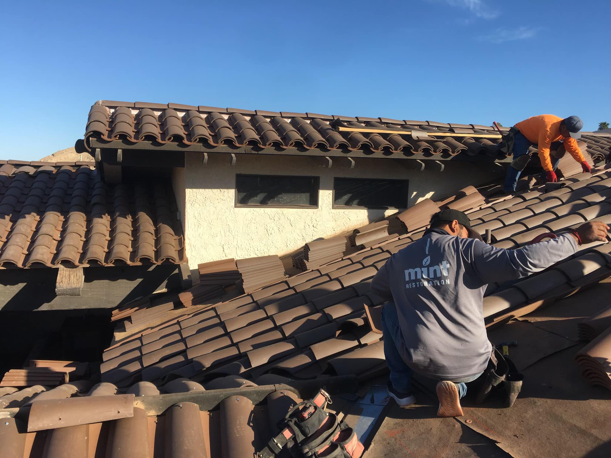 Two workers repairing a damaged tiled roof with tools and materials around them.