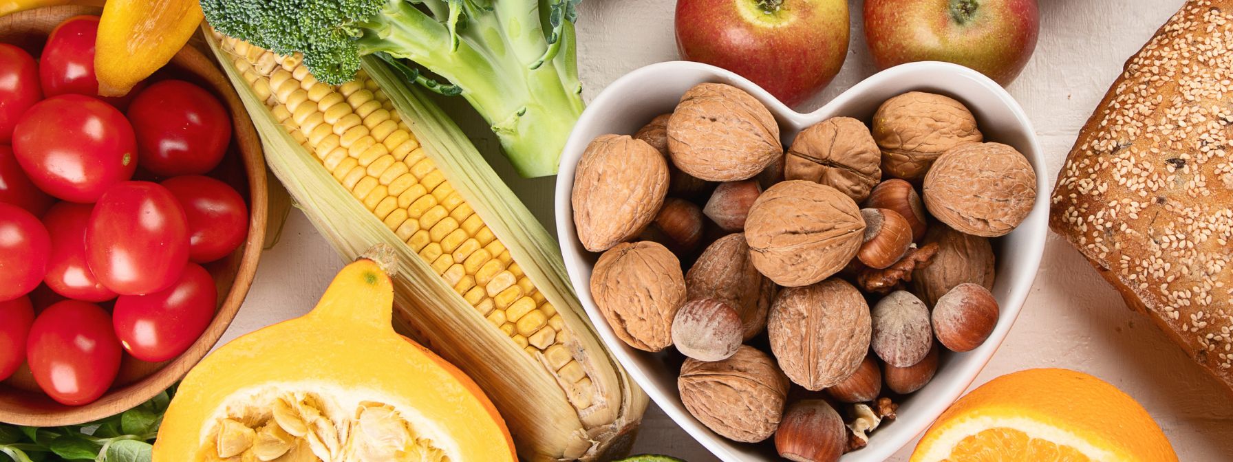 An assortment of whole foods including cherry tomatoes, corn on the cob, broccoli, apples, mixed nuts in a heart-shaped bowl, whole grain bread, squash, and orange slices arranged on a light surface. An assortment of whole foods including cherry tomatoes, corn on the cob, broccoli, apples, mixed nuts in a heart-shaped bowl, whole grain bread, squash, and orange slices arranged on a light surface.