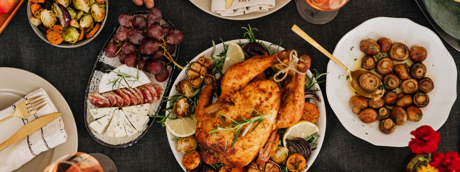 A whole roasted chicken served with lemon, garlic, and vegetables, surrounded by plates of sautéed mushrooms, roasted Brussels sprouts, grapes, cheese, and salami on a festive dinner table. A whole roasted chicken served with lemon, garlic, and vegetables, surrounded by plates of sautéed mushrooms, roasted Brussels sprouts, grapes, cheese, and salami on a festive dinner table.