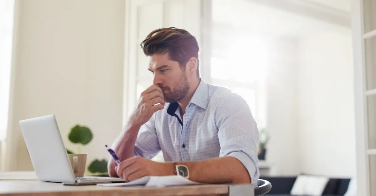 A man sitting at a desk with a laptop, holding a pen and looking thoughtful while working. A man sitting at a desk with a laptop, holding a pen and looking thoughtful while working.