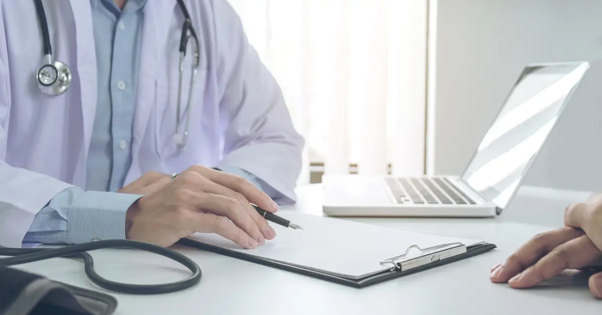 A doctor with a stethoscope sits at a desk holding a pen over a clipboard, with a laptop and another person’s hand visible on the table. A doctor with a stethoscope sits at a desk holding a pen over a clipboard, with a laptop and another person’s hand visible on the table.