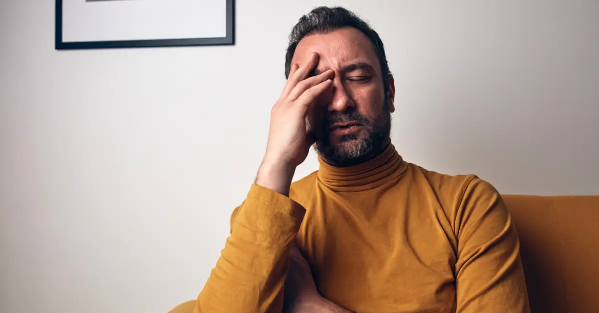 A middle-aged man in a mustard turtleneck sits with his eyes closed, holding his forehead with a pained or stressed expression.