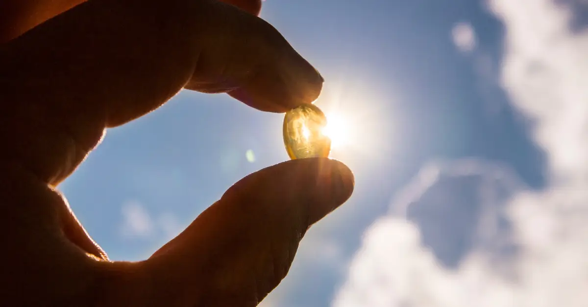 Close-up of a hand holding a translucent supplement capsule against the bright sky and sunlight.