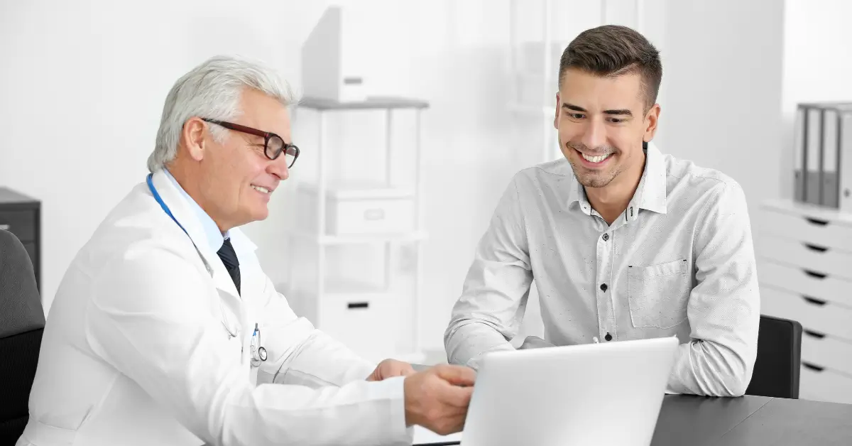 A doctor in a white coat sits beside a smiling patient, both looking at a laptop screen during a consultation in a bright, modern medical office.