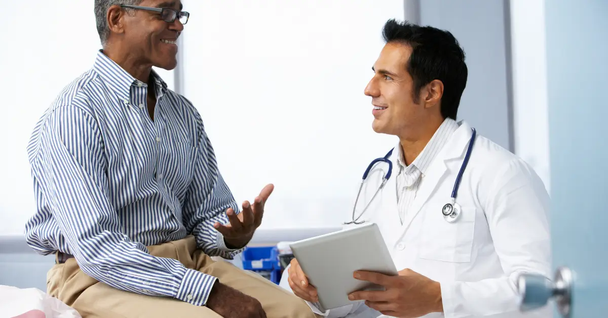 An older man sitting on an exam table talks and smiles with a doctor in a white coat holding a digital tablet, during a medical consultation in a bright clinic room. An older man sitting on an exam table talks and smiles with a doctor in a white coat holding a digital tablet, during a medical consultation in a bright clinic room.