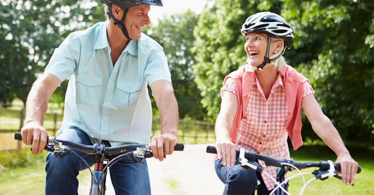 A man and woman ride bicycles outdoors on a sunny day, smiling at each other while wearing helmets as they cycle along a tree-lined path. A man and woman ride bicycles outdoors on a sunny day, smiling at each other while wearing helmets as they cycle along a tree-lined path.