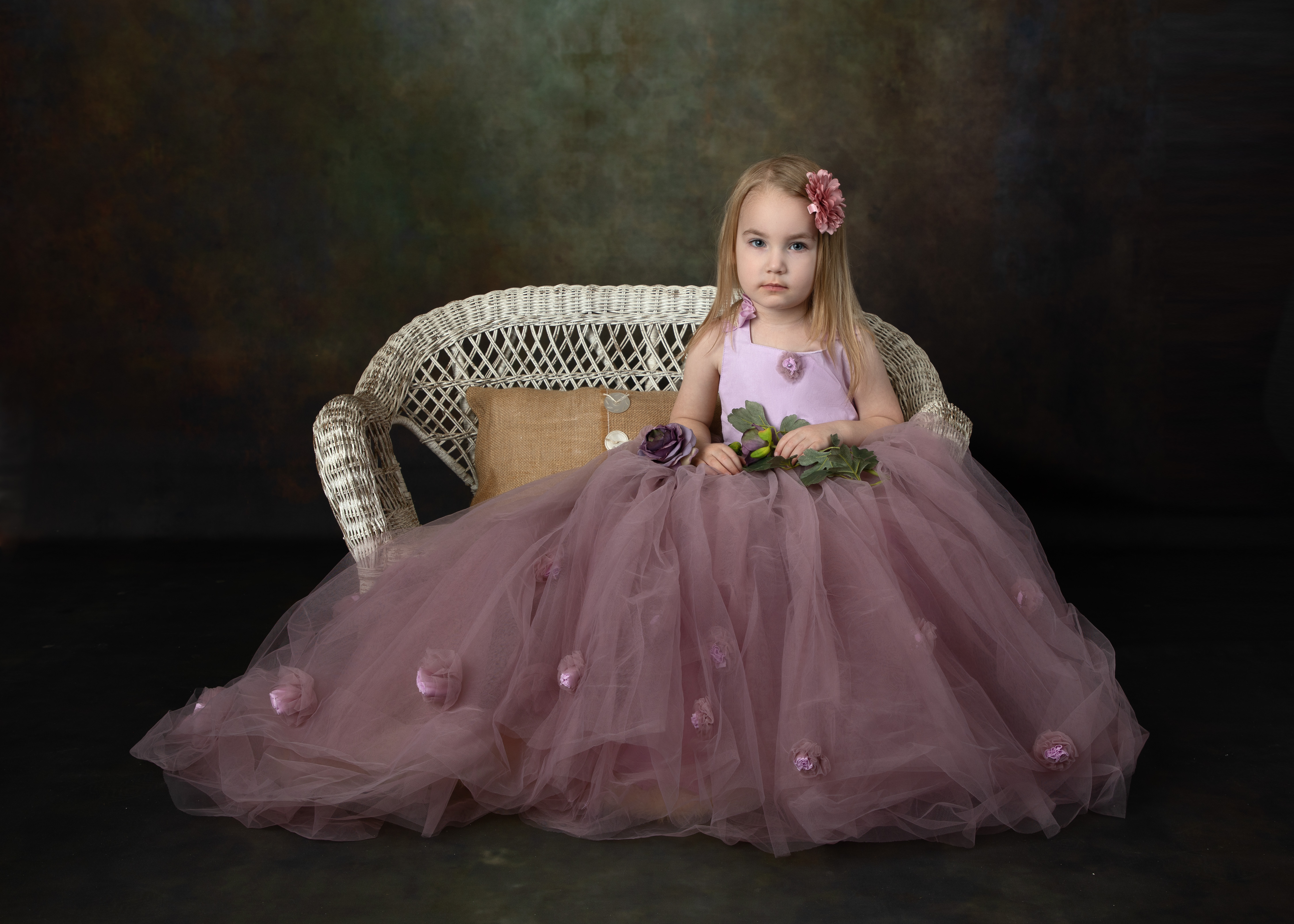 Formal portrait of a little girl in a long purple dress sitting on a white wicker bench. 