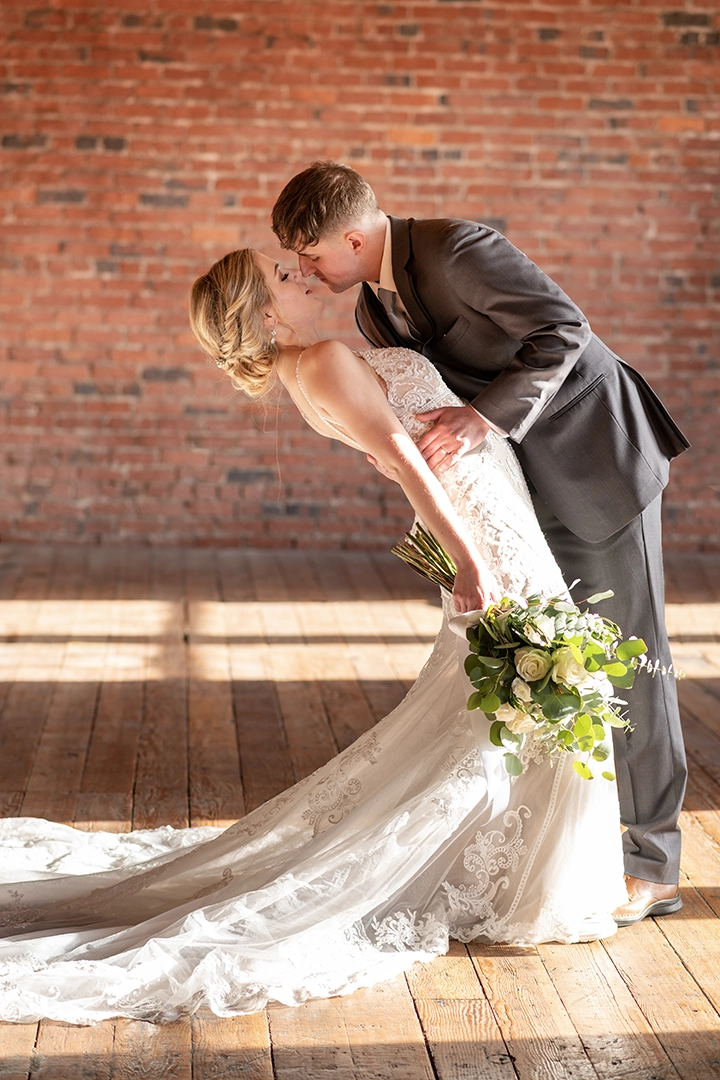Bride and groom share a romantic kiss as the groom dips the bride in a sunlit PNW warehouse with exposed brick walls and wooden floors. The bride holds a white and green bouquet, and her lace train flows across the floor. Captured by a wedding photographer in Portland.