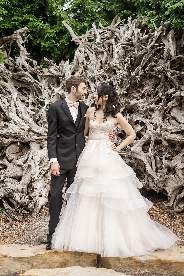 Bride and groom standing arm-in-arm in front of a dramatic, textured driftwood backdrop, sharing an intimate gaze. The bride wears a layered tulle gown with floral appliqué, and the groom is in a classic black tuxedo. A stunning example of Seattle wedding photos that blend natural elegance with editorial sophistication.