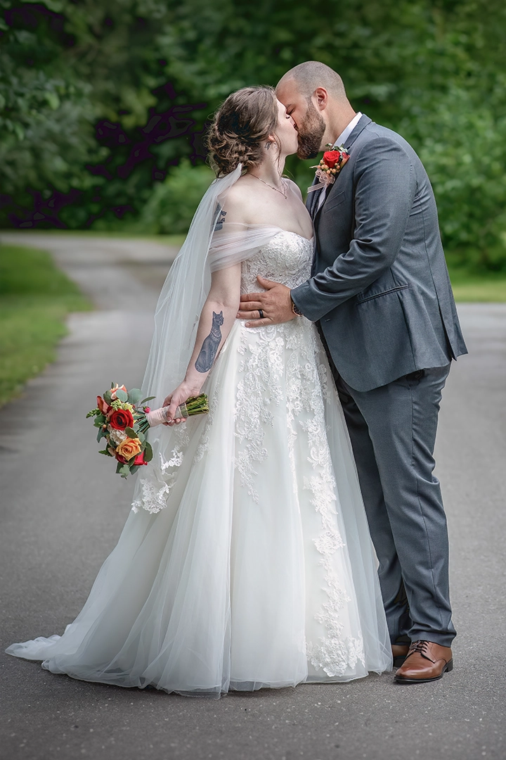 Bride and groom share a romantic kiss on a quiet tree-lined path, with the bride holding a bouquet of coral and red flowers and wearing a lace wedding gown with a sheer veil. Captured during their elegant summer celebration—fine wedding photography in Maple Valley, WA.