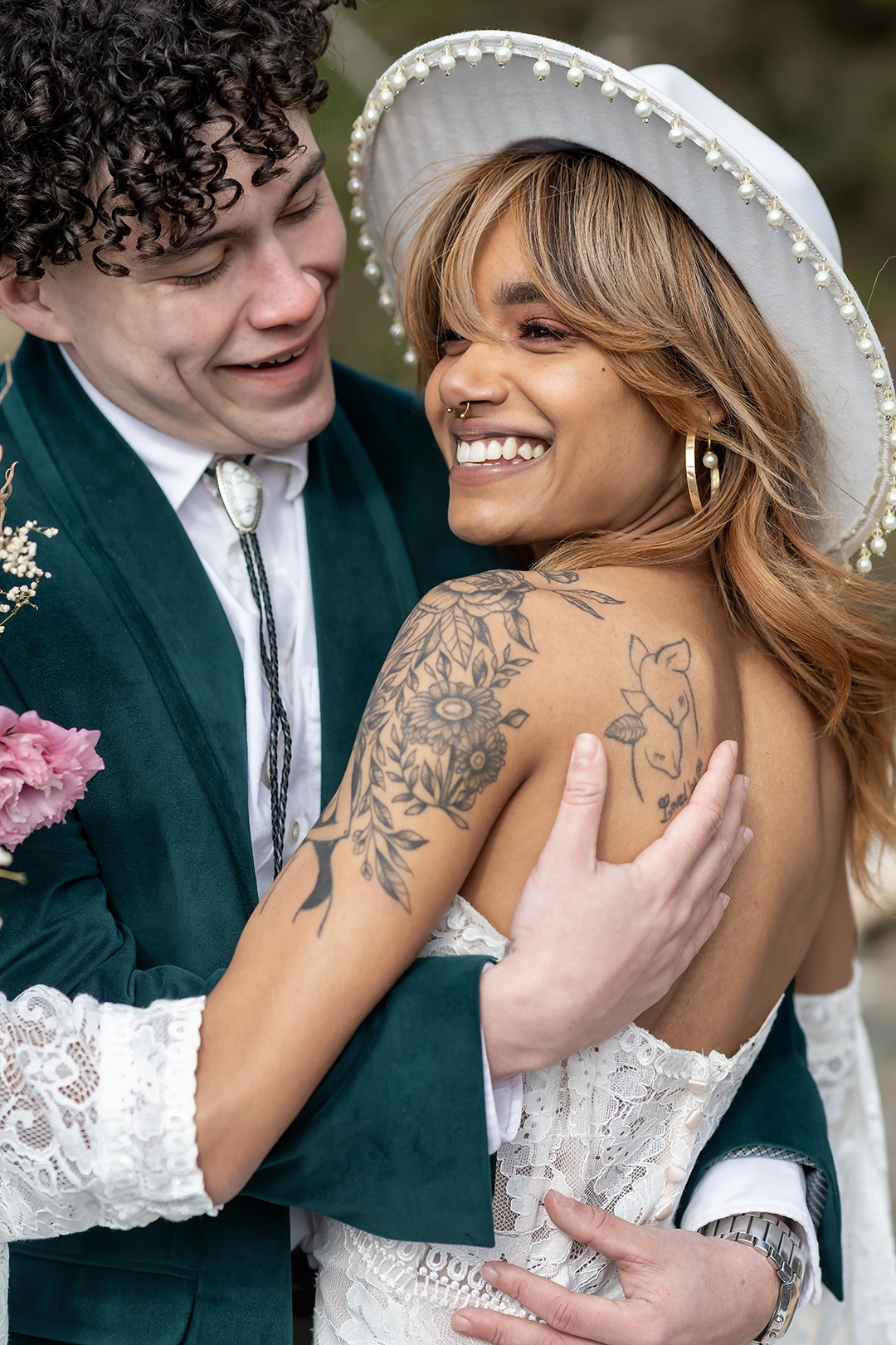 A joyful bride and groom embrace during their Cannon Beach wedding. The bride, glowing with a wide smile, wears a white hat trimmed with pearls and an off-the-shoulder lace gown, revealing beautifully detailed tattoos. The groom, smiling at her with pure admiration, is dressed in a deep green velvet suit and bolo tie. Their connection is effortless, stylish, and full of warmth.
