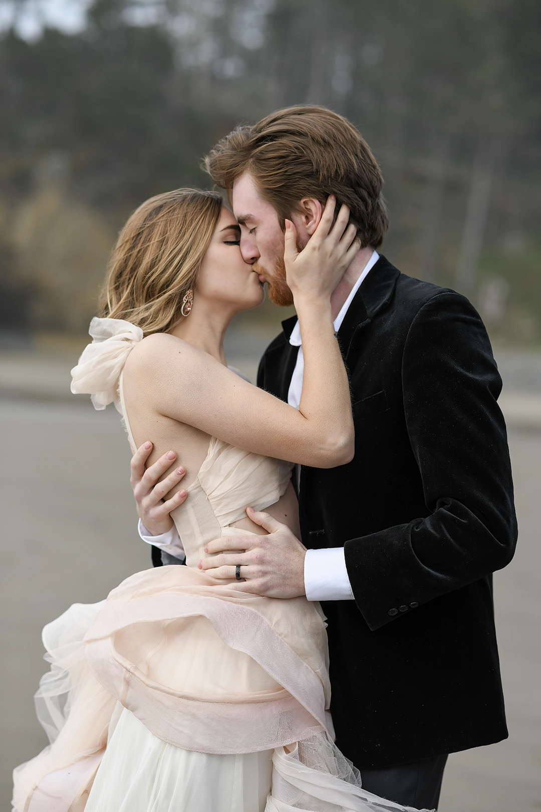 Kelsey and Braxton share a deeply romantic kiss along the Oregon Coast. The bride’s blush-toned, ethereal gown sways in soft layers as she gently holds her groom’s face. He wears a classic black velvet jacket, embracing her with timeless devotion. The moment is raw, elegant, and cinematic—capturing the essence of modern romance against a muted coastal backdrop.