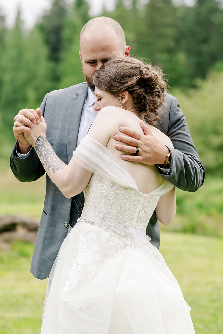 A tender moment between a bride and groom during their outdoor wedding near Seattle. The groom embraces the bride lovingly as they share a quiet dance surrounded by lush greenery. The bride’s lace gown and the groom’s tailored suit highlight the elegance and intimacy of this candid wedding portrait captured by Seattle wedding photographer, Adina Stiles Photography.