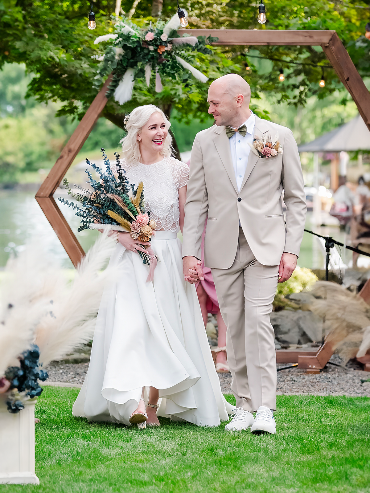 A chic bride in a lace gown and veil, holding a bouquet of wild blooms, walks hand-in-hand with her dapper groom under a wooden arch, embodying the refined elegance of a Pacific Northwest wedding celebration.