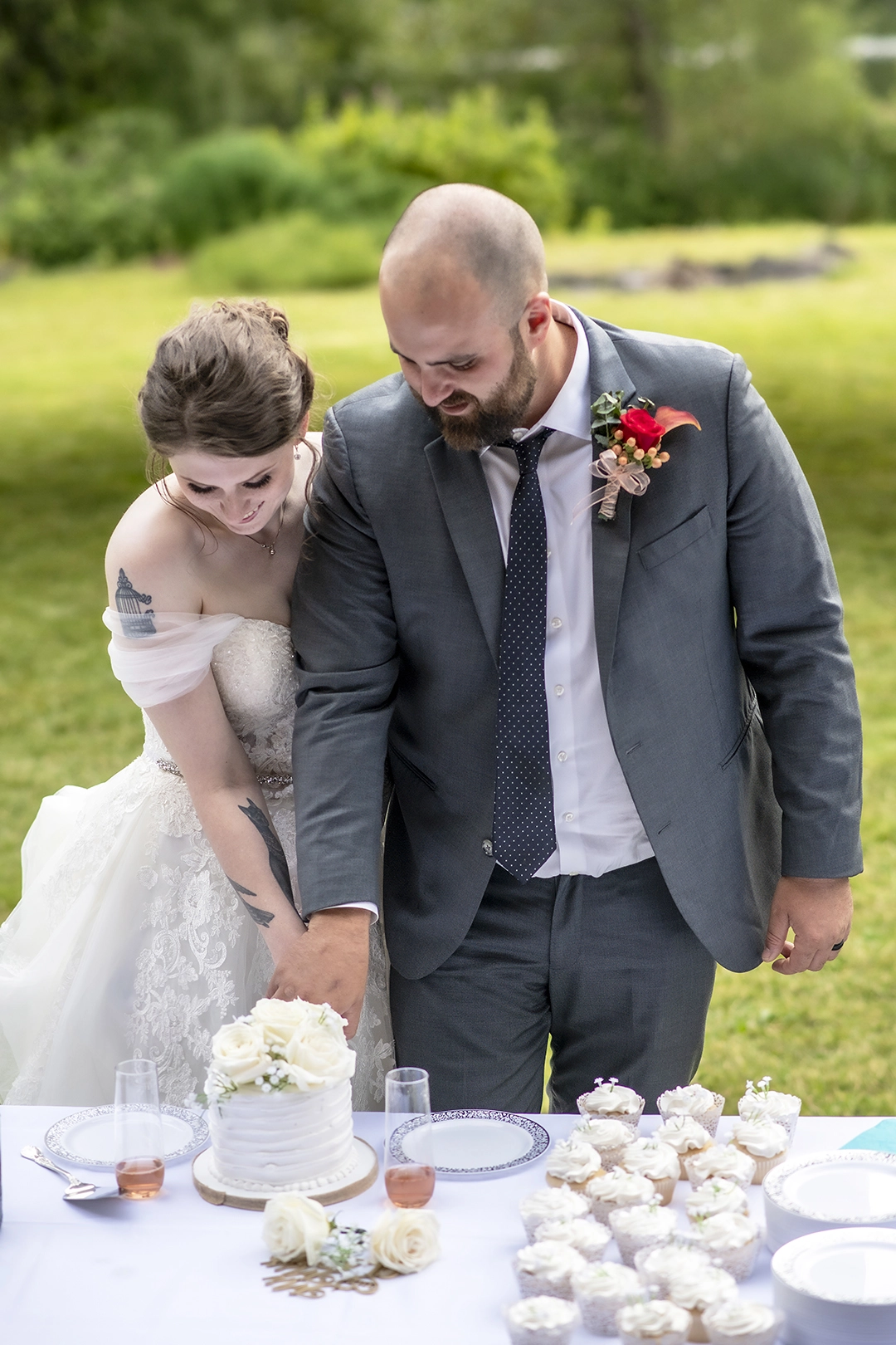 A sophisticated couple in timeless attire cuts into an elegant white cake adorned with roses, surrounded by delicate cupcakes, amidst a lush green outdoor setting—capturing the essence of a Seattle wedding photographer's artistry in crafting enduring love stories.