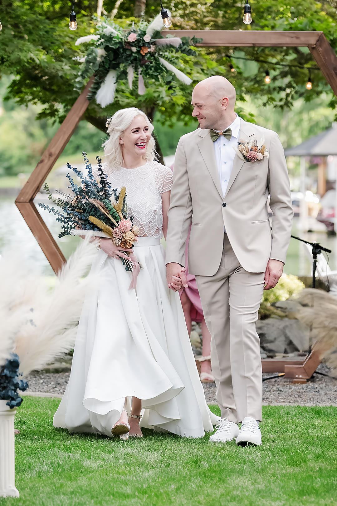 A joyful bride and groom share an intimate forehead-to-forehead moment by the lake, radiating love and elegance. Her pearl-draped top and softly gathered skirt shimmer with quiet luxury, while his tailored beige suit adds timeless refinement. Captured by wedding photographer Adina Stiles, this image evokes the polished intimacy and enduring style of a true heirloom.