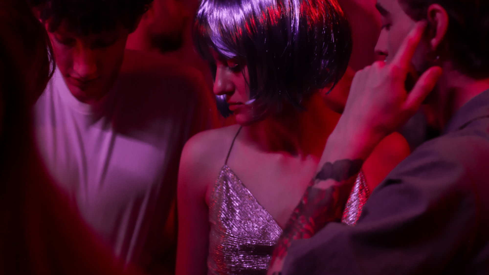 Woman in a purple bobbed wig wearing a sparkling strapped dress with bare arms and shoulders, looking to her right. Dark haired caucasian male in white tshirt looking down to her right and to her left a dark-haired man with his finger in his ear.