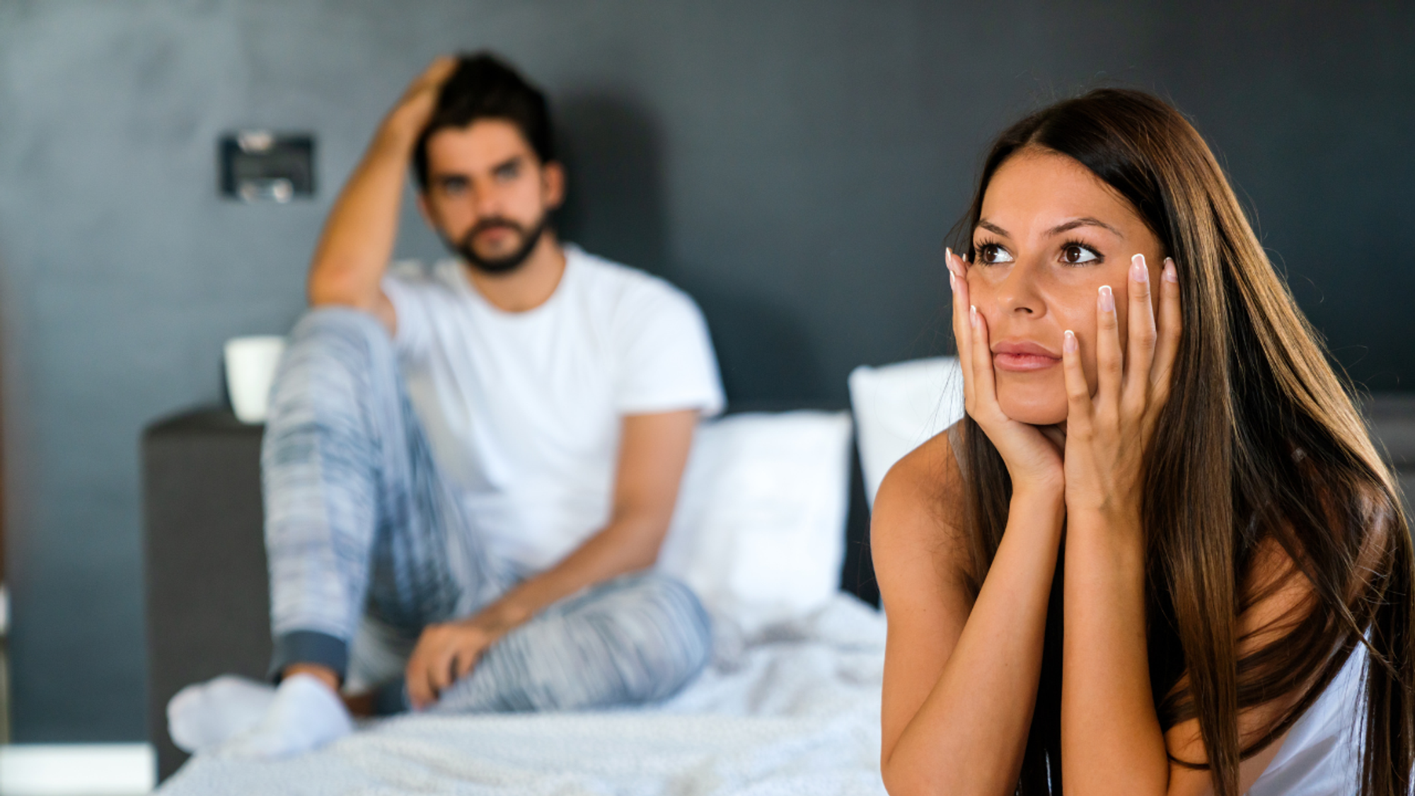 Caucasian man and woman couple on bed, brunette lady in the foreground looks troubled