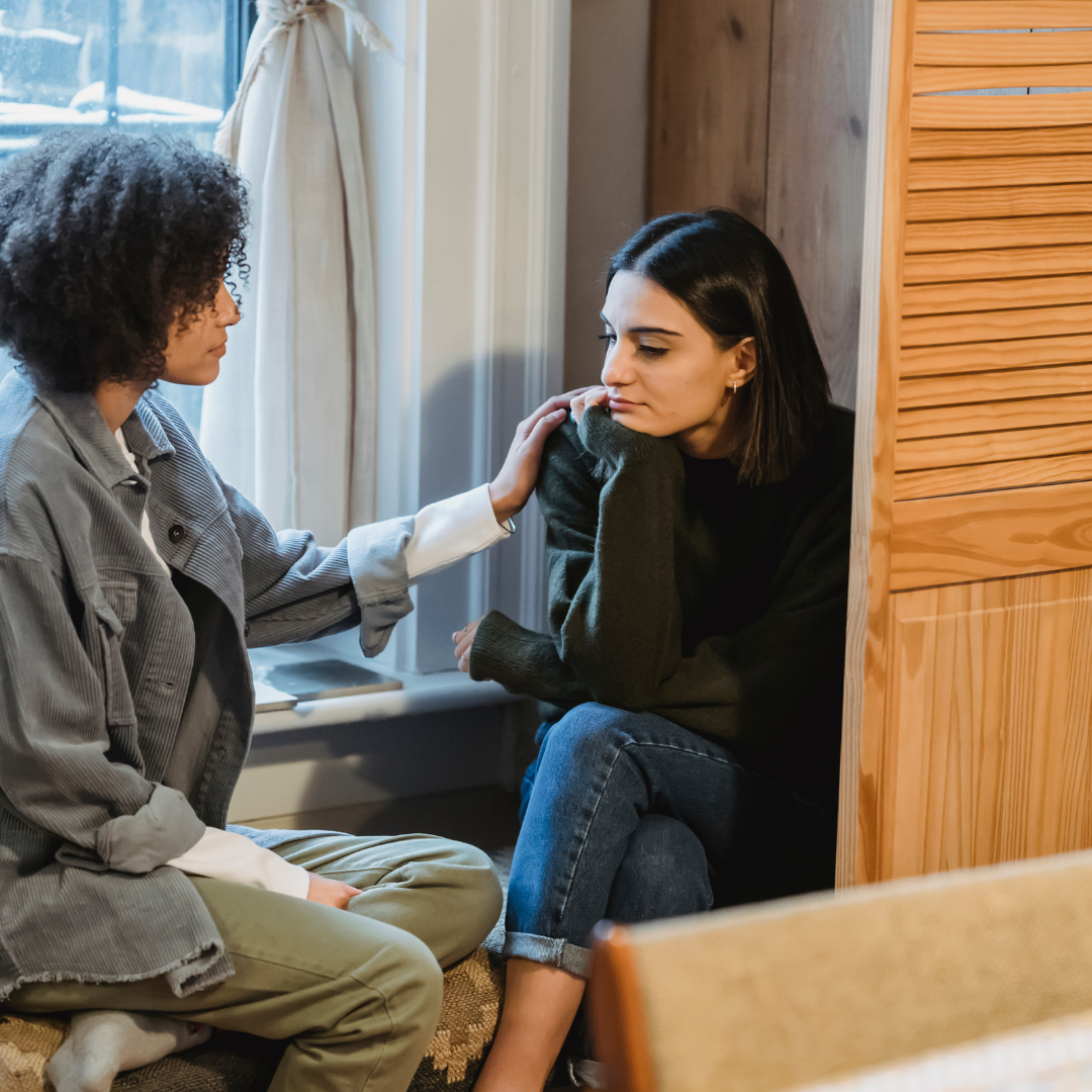 Two women sitting by a window, one offering a comforting hand on the other's shoulder during an emotional conversation, illustrating the power of personal support through holistic medicine.