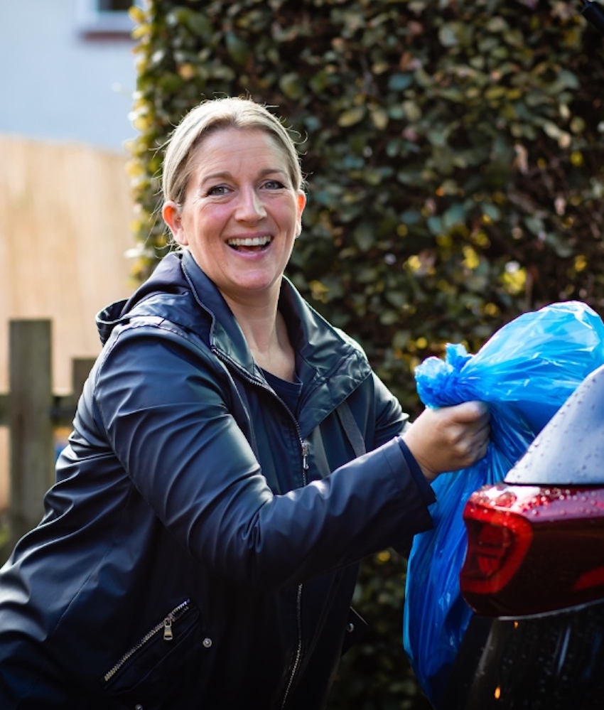 Smiling woman bundling large bin bags into the boot of a car.