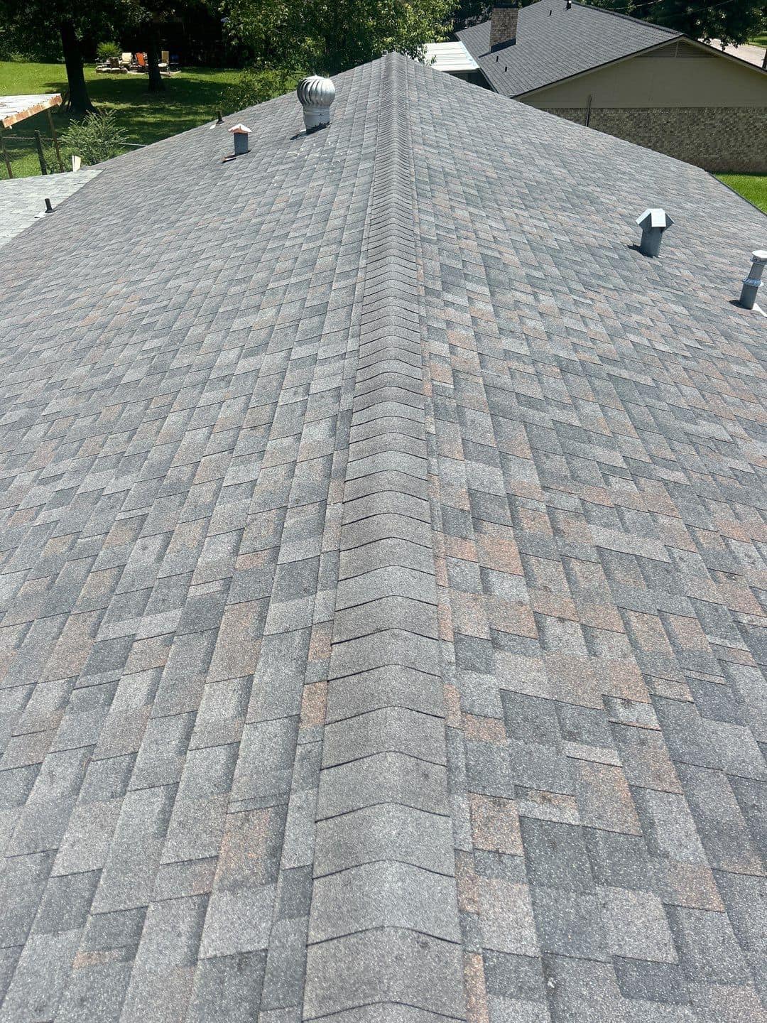 A close-up view of a roof with grey asphalt shingles, showing significant hail damage. The shingles display numerous dents and bruises indicative of hail impact. The roof is equipped with multiple vents and a turbine vent. The surrounding area includes neighboring rooftops and greenery, indicating a residential neighborhood. The damage is extensive, affecting the uniform appearance of the shingles. A close-up view of a roof with grey asphalt shingles, showing significant hail damage. The shingles display numerous dents and bruises indicative of hail impact. The roof is equipped with multiple vents and a turbine vent. The surrounding area includes neighboring rooftops and greenery, indicating a residential neighborhood. The damage is extensive, affecting the uniform appearance of the shingles.
