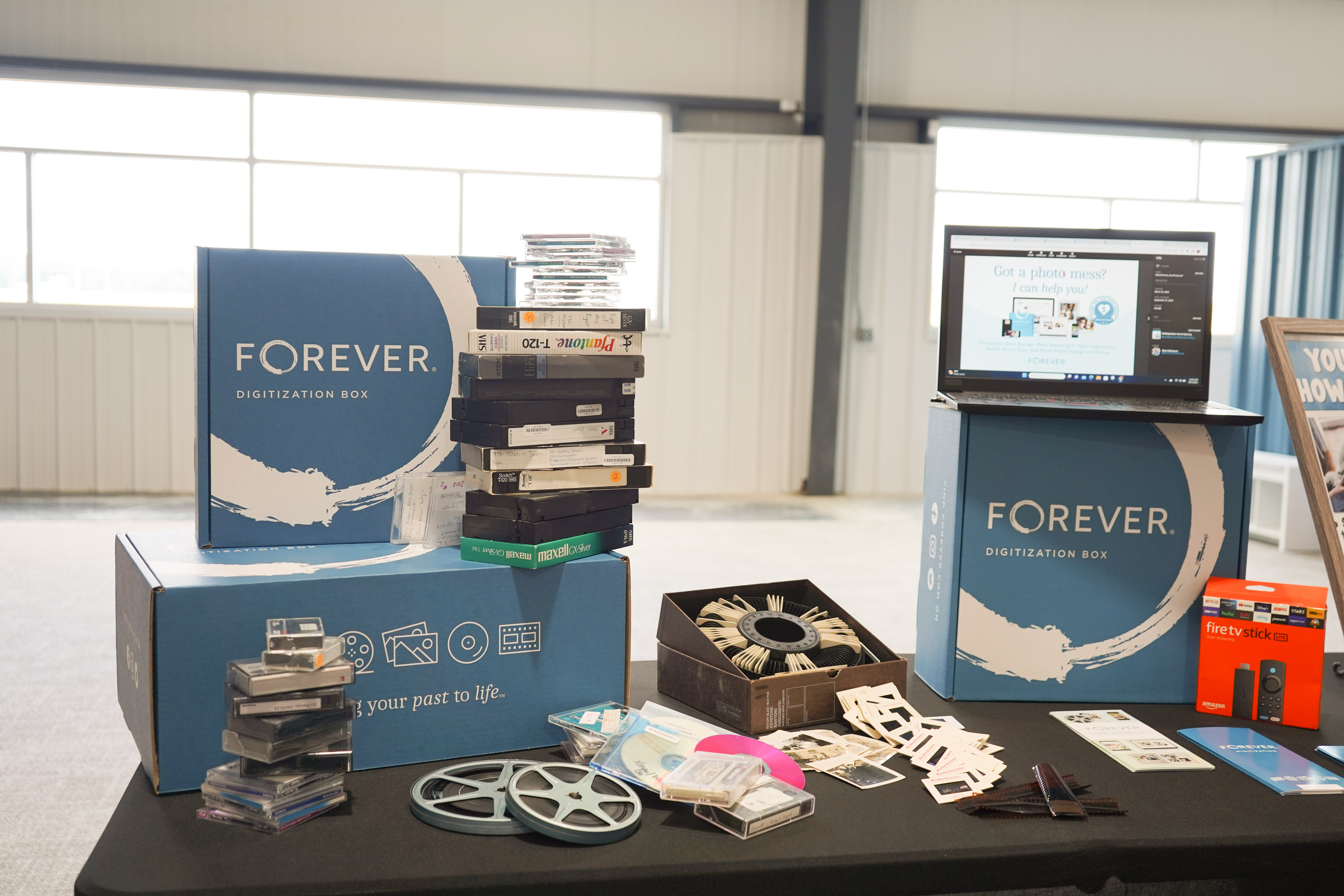 Old videos, reel to reel film, piled up on a table with old photos and a FOREVER digitization box nearby. Old videos, reel to reel film, piled up on a table with old photos and a FOREVER digitization box nearby.