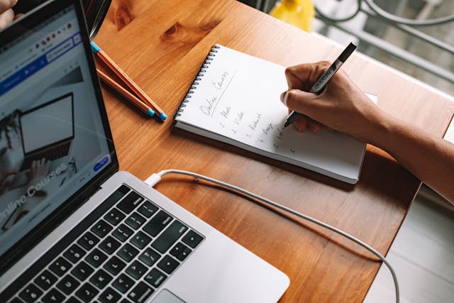 Person writing on a notepad beside a laptop Person writing on a notepad beside a laptop
