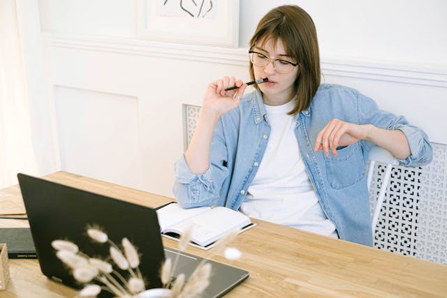 Woman holding pen near mouth while looking at her notebook on the table Woman holding pen near mouth while looking at her notebook on the table