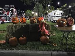 Nighttime outdoor Halloween display featuring carved pumpkins on hay bales and tables at a community Trunk or Treat event, with fire trucks and festive lights in the background. Nighttime outdoor Halloween display featuring carved pumpkins on hay bales and tables at a community Trunk or Treat event, with fire trucks and festive lights in the background.