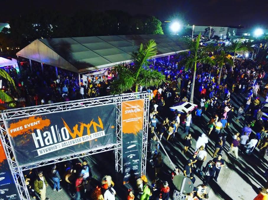 Aerial nighttime view of Wynwood’s HalloWYN Block Party in Miami showing large crowds, palm trees, and a brightly lit event entrance banner reading ‘HalloWYN – Wynwood’s Halloween Block Party. Aerial nighttime view of Wynwood’s HalloWYN Block Party in Miami showing large crowds, palm trees, and a brightly lit event entrance banner reading ‘HalloWYN – Wynwood’s Halloween Block Party.