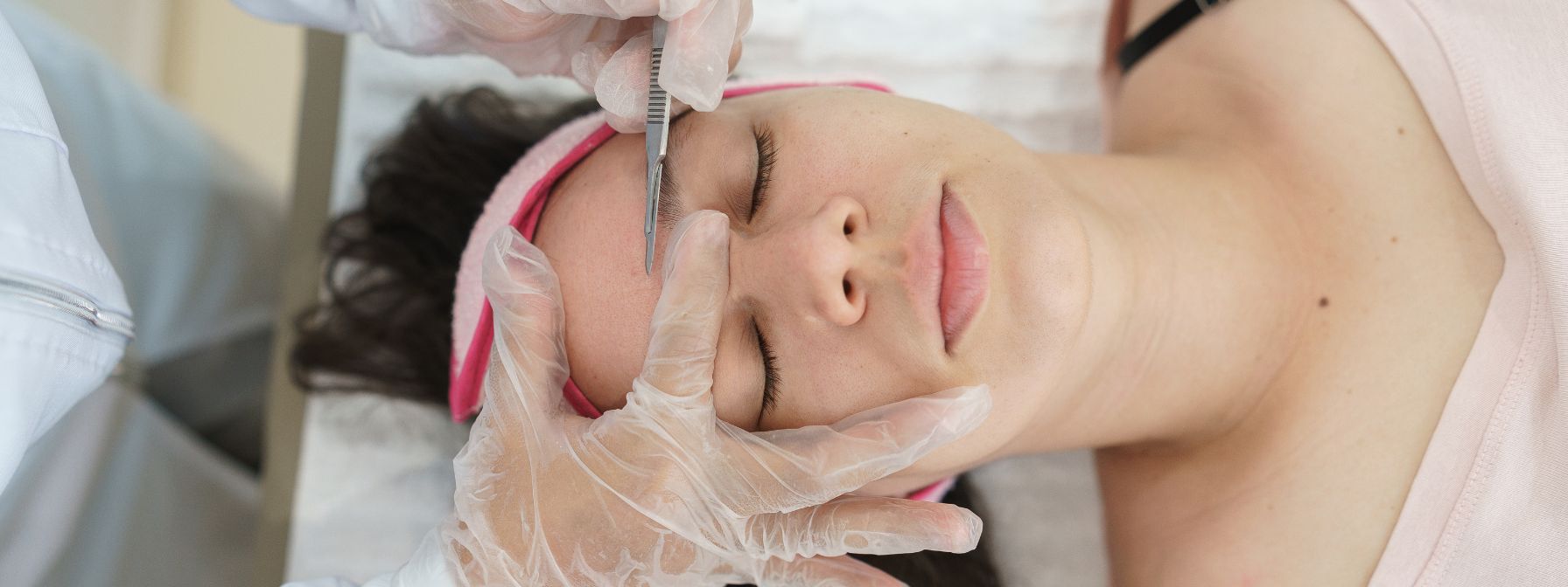 A close-up of a woman receiving a professional dermaplaning treatment, as a skincare specialist uses a sterile scalpel to gently exfoliate her forehead while she rests with eyes closed. A close-up of a woman receiving a professional dermaplaning treatment, as a skincare specialist uses a sterile scalpel to gently exfoliate her forehead while she rests with eyes closed.