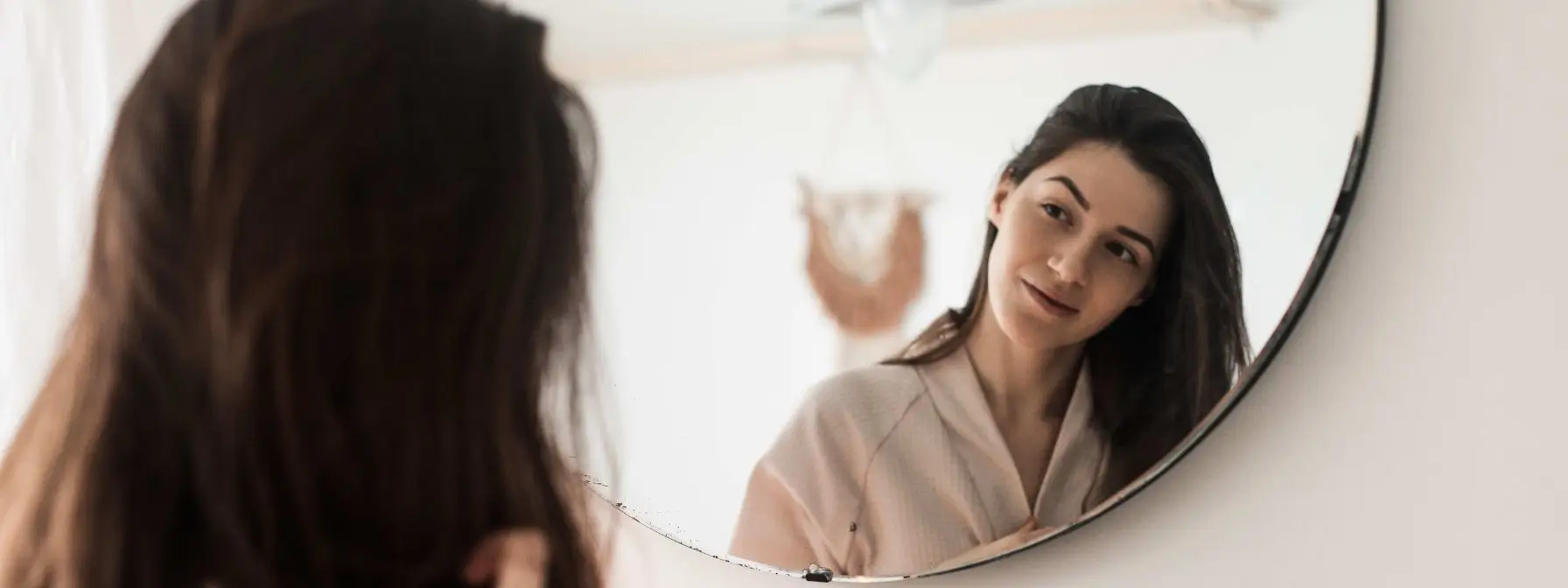 A woman wearing a robe smiles at her reflection in a round mirror, showing clear, smooth skin in a softly lit, minimalist bathroom setting. A woman wearing a robe smiles at her reflection in a round mirror, showing clear, smooth skin in a softly lit, minimalist bathroom setting.