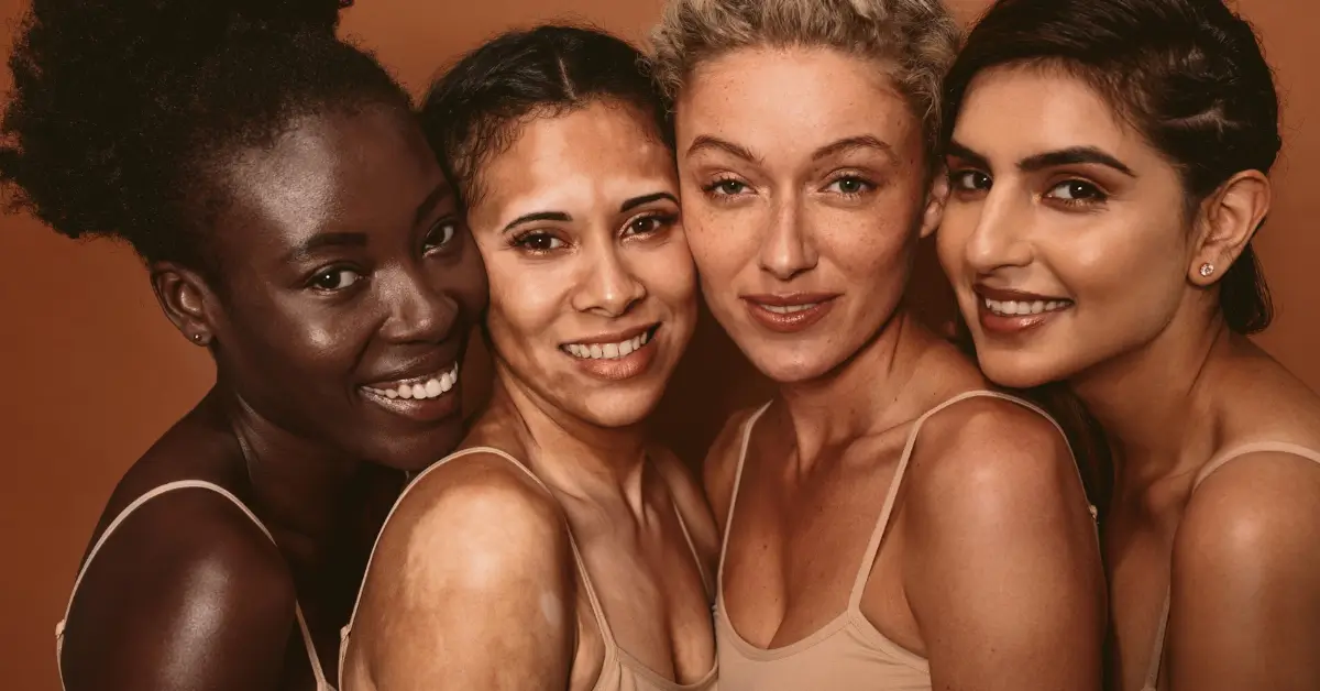 Four women with diverse skin tones smiling closely together against a warm brown background, showcasing healthy, radiant skin as a result of personalized skincare routines.