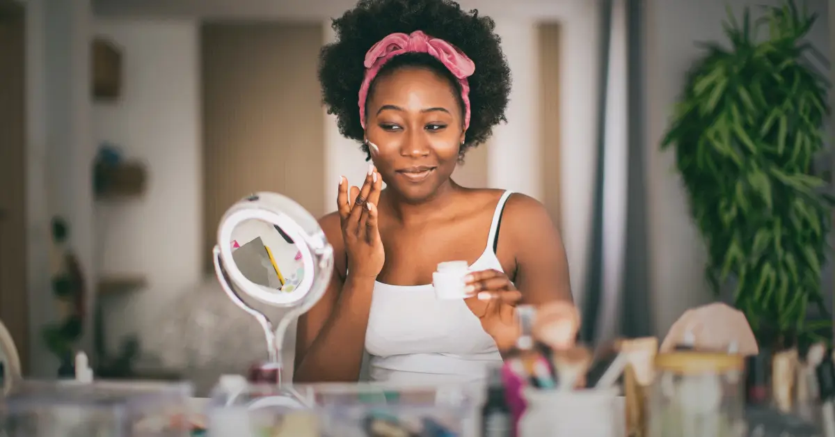 Woman smiling while applying face cream in front of a mirror, surrounded by skincare and beauty products, illustrating a personalized skincare routine for glowing skin in Greenville, SC.