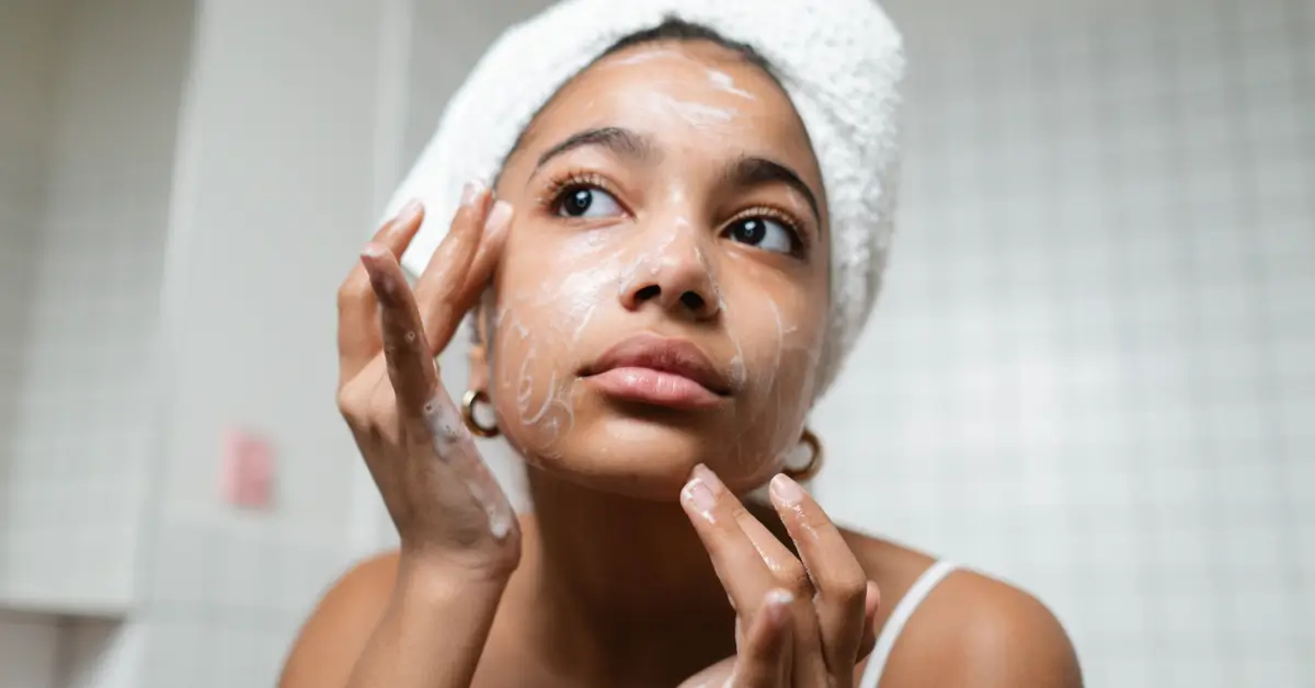Young woman with a towel wrapped around her head applying a foaming cleanser to her face