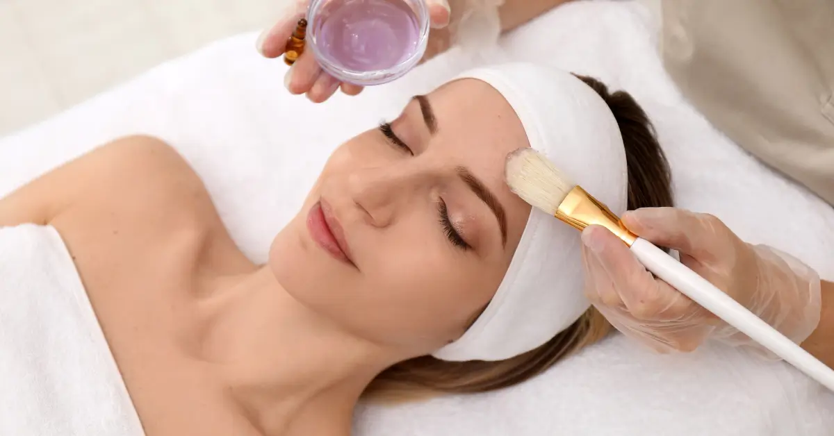 Woman with eyes closed receiving a chemical peel treatment as an aesthetician applies a purple solution to her forehead with a brush, Woman with eyes closed receiving a chemical peel treatment as an aesthetician applies a purple solution to her forehead with a brush,