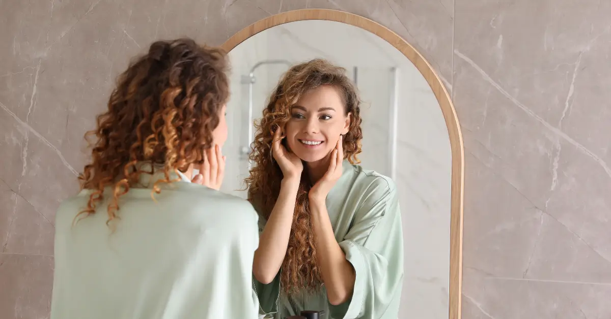 Smiling woman in a robe gently touching her face while admiring her glowing skin in the bathroom mirror Smiling woman in a robe gently touching her face while admiring her glowing skin in the bathroom mirror