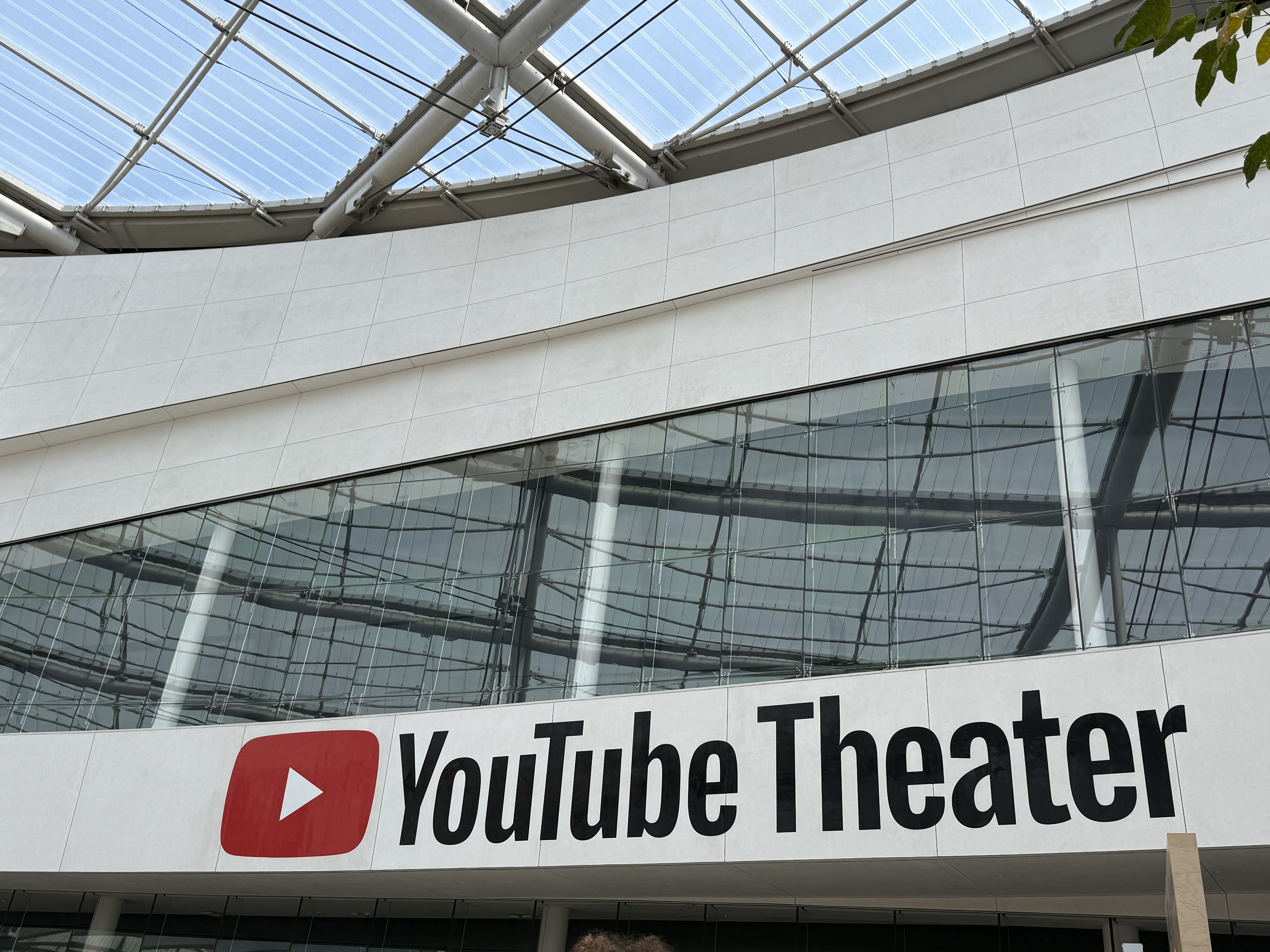 Exterior view of YouTube Theater with large red and white YouTube logo and bold black lettering on the building facade, under a glass roof structure.