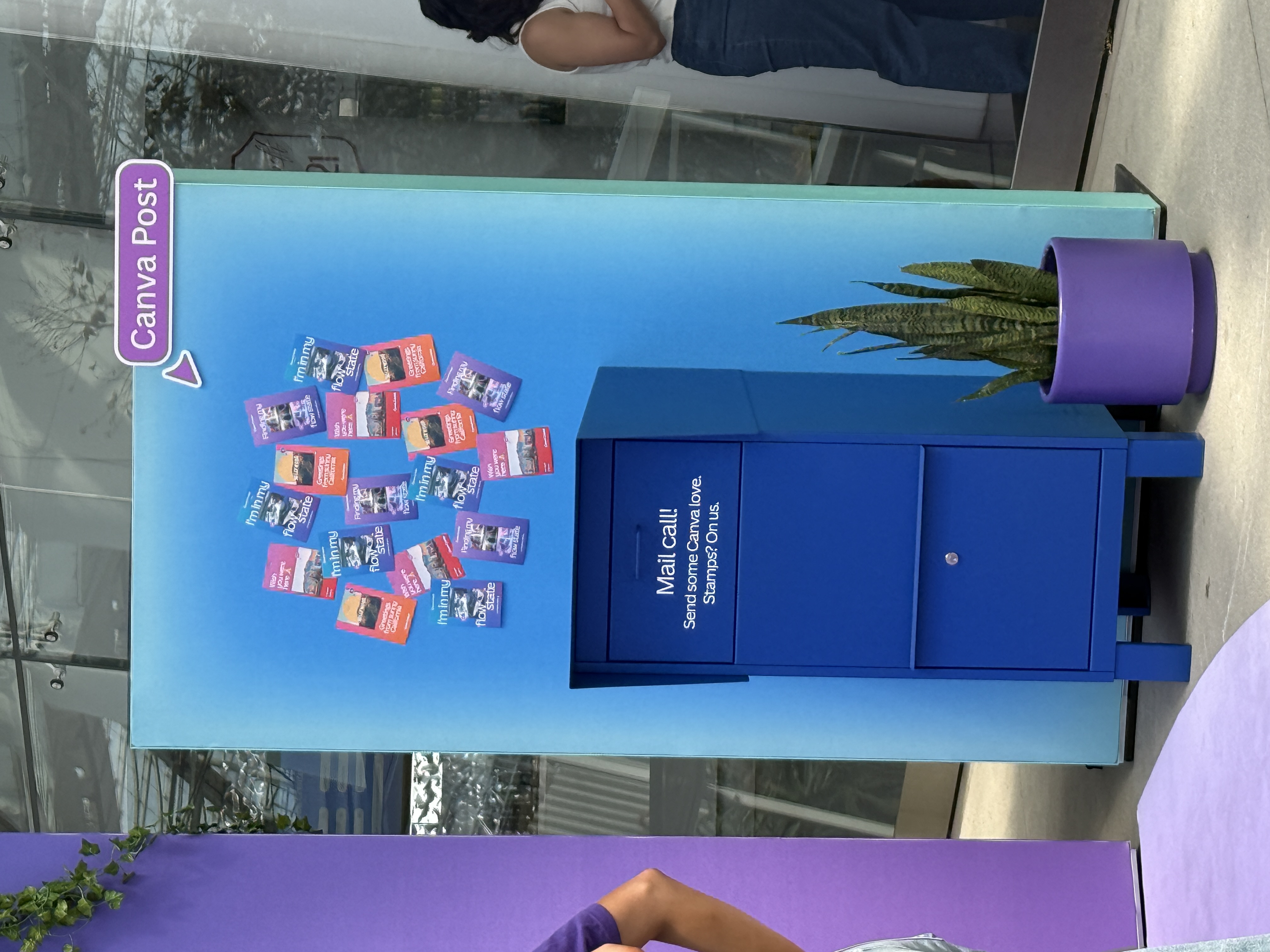 Bright blue mailbox labeled ‘Mail Call! Send some Canva love. Stamps? On us.’ at Canva Create, with colorful postcards displayed on a gradient backdrop and a potted plant beside it.