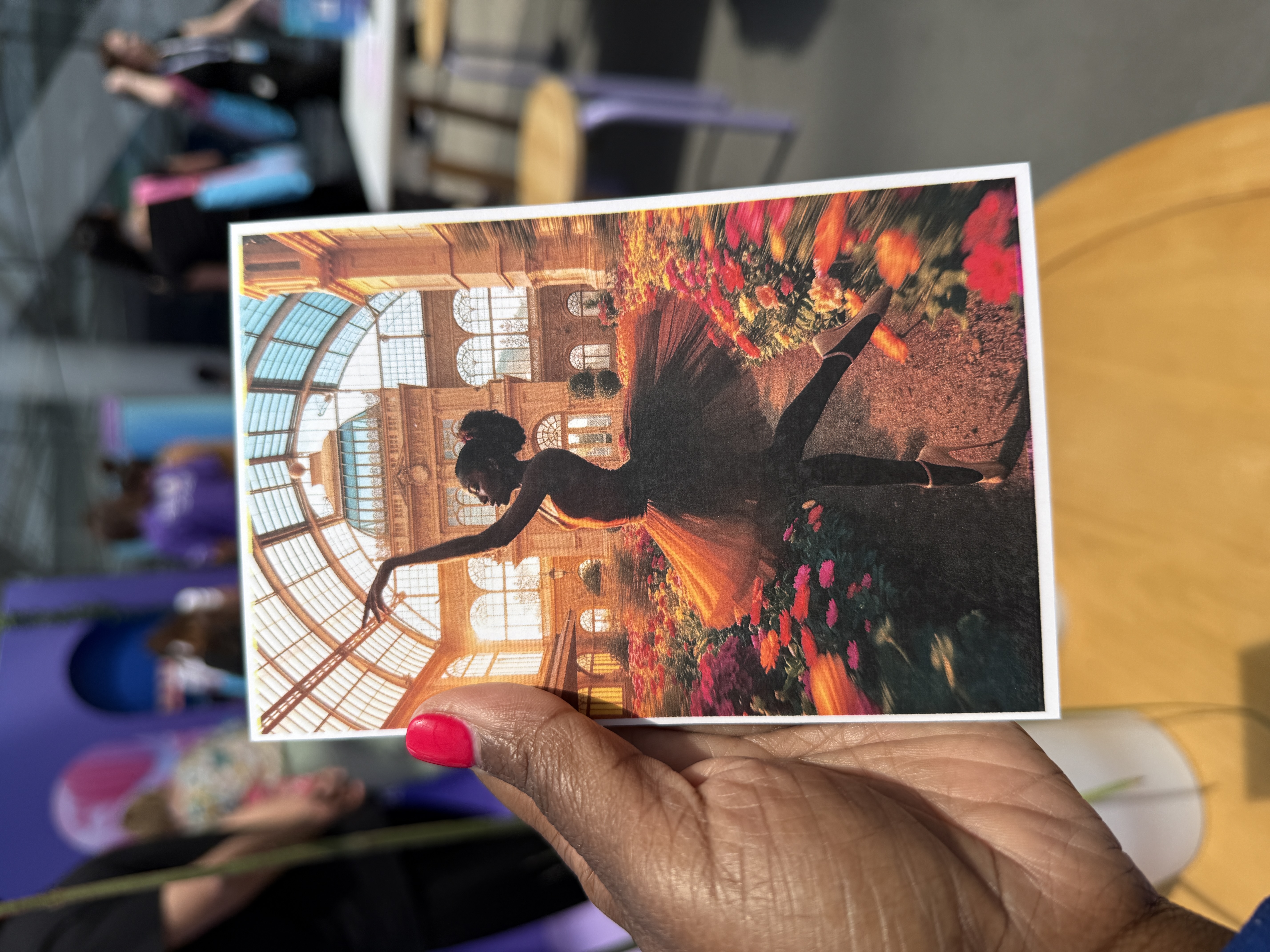 Hand holding a postcard created at Canva Create, featuring an artistic image of a ballerina silhouetted against a sunlit glass atrium with vibrant flowers in the foreground.”