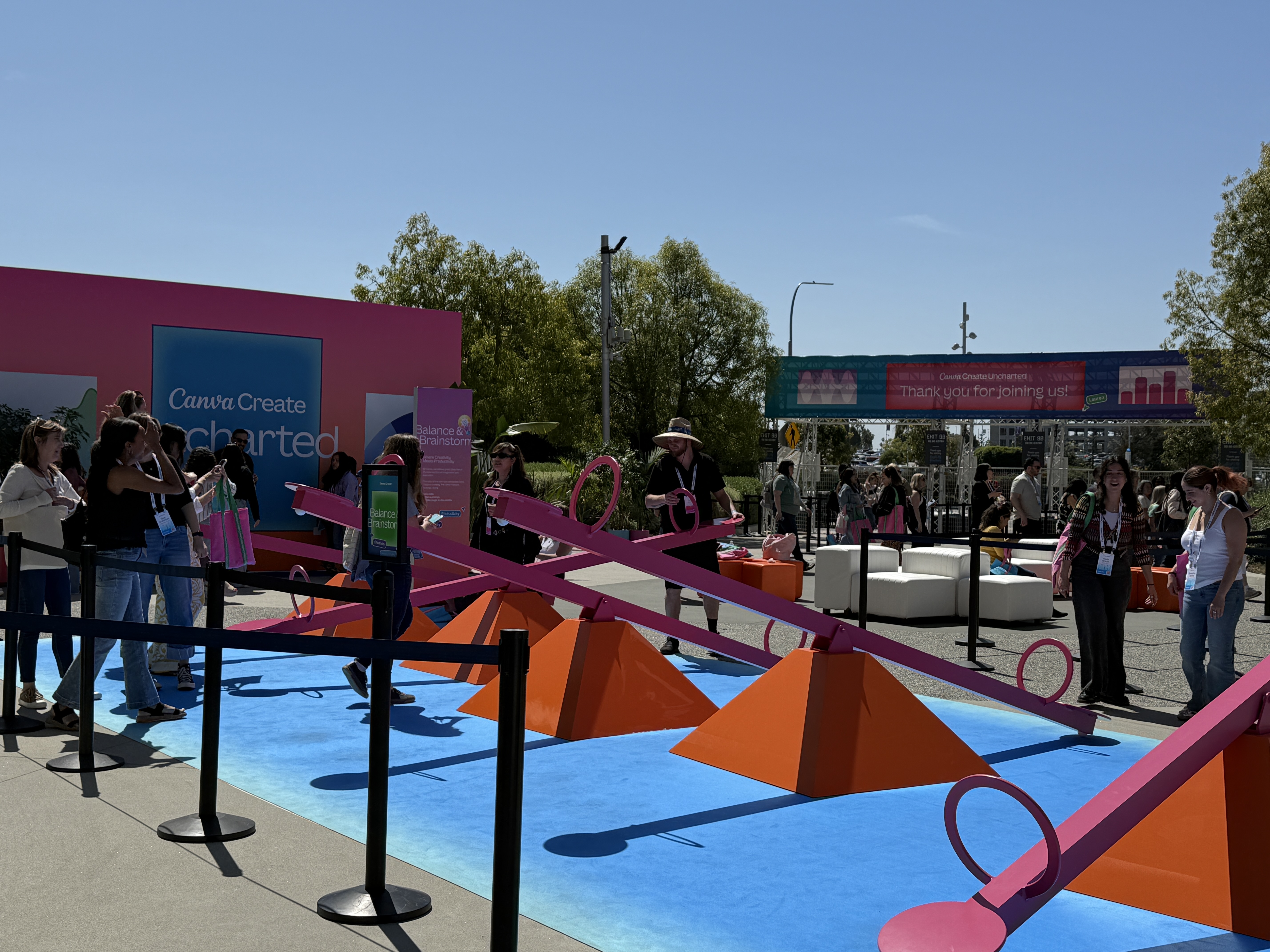Outdoor play area at Canva Create featuring bright pink seesaws on orange bases, with attendees participating and watching under a sunny sky, surrounded by colorful event signage.