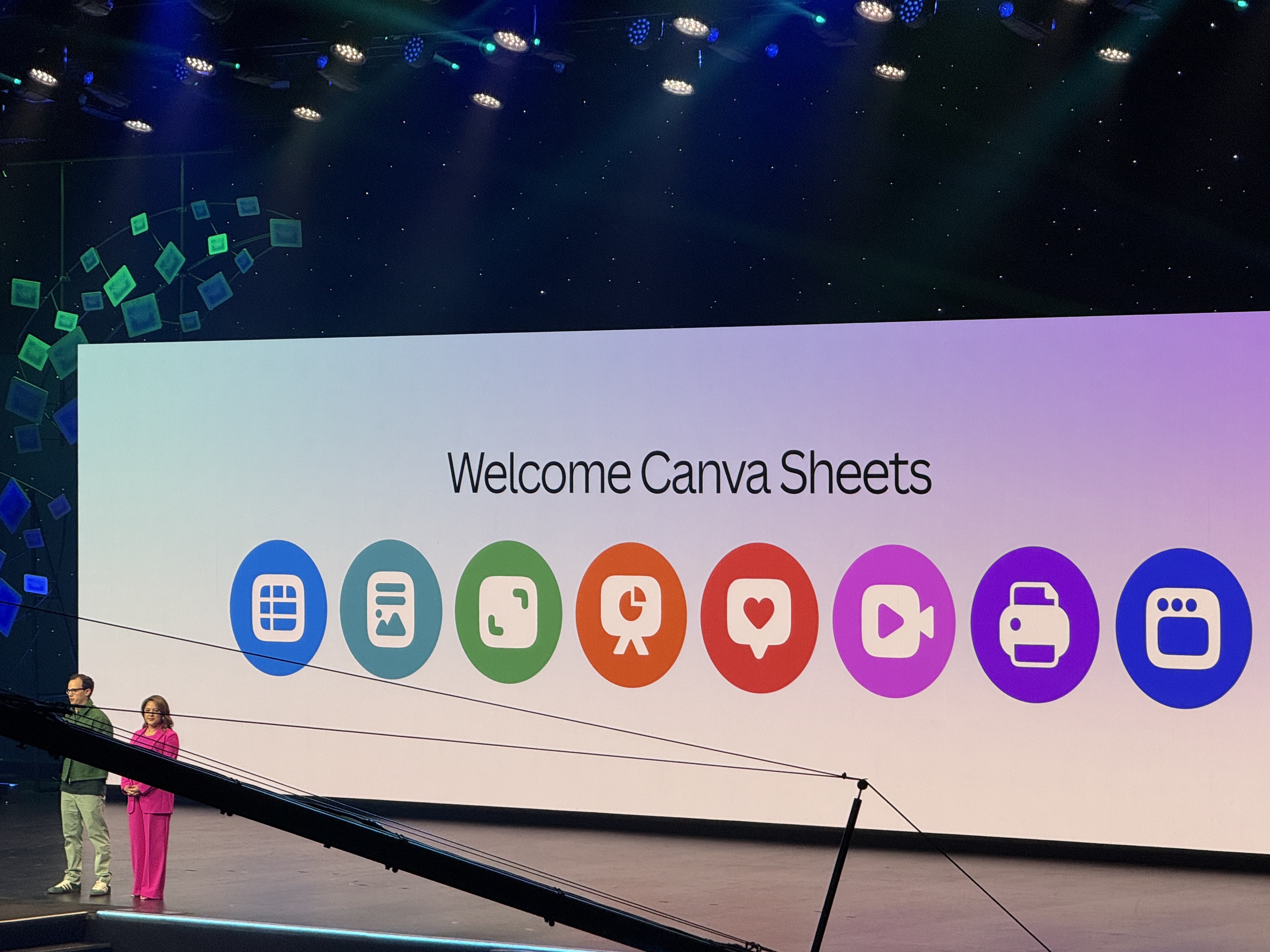 Conference stage at Canva Create showing a large screen with the words ‘Welcome Canva Sheets’ and colorful icons representing different Canva tools, with two presenters standing at the left.